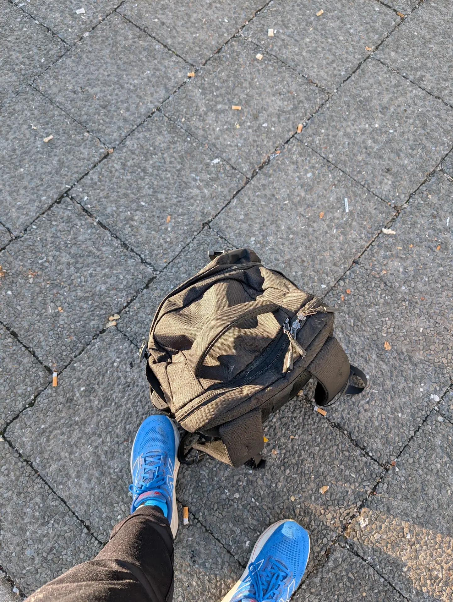Ein brauner Rucksack steht zwischen blauen Laufschuhen auf dem Pflaster.