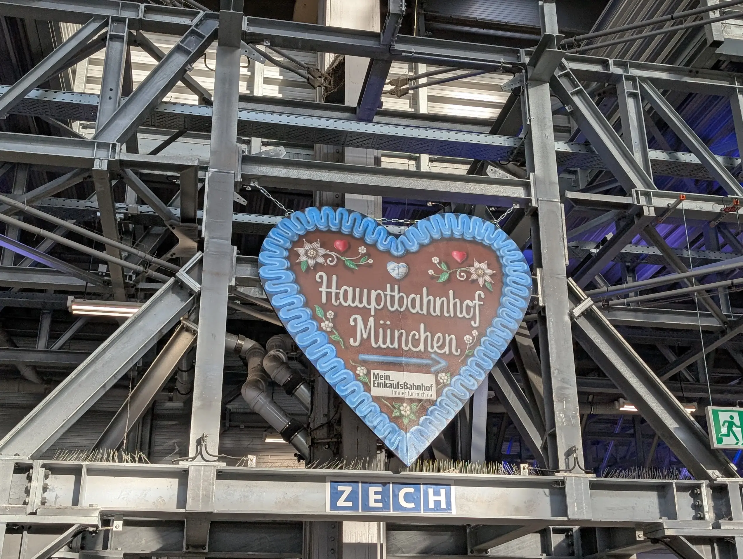Lebkuchenherz mit der Aufschrift „Hauptbahnhof München“ vor der sichtbaren Stahlkonstruktion des Bahnhofs.