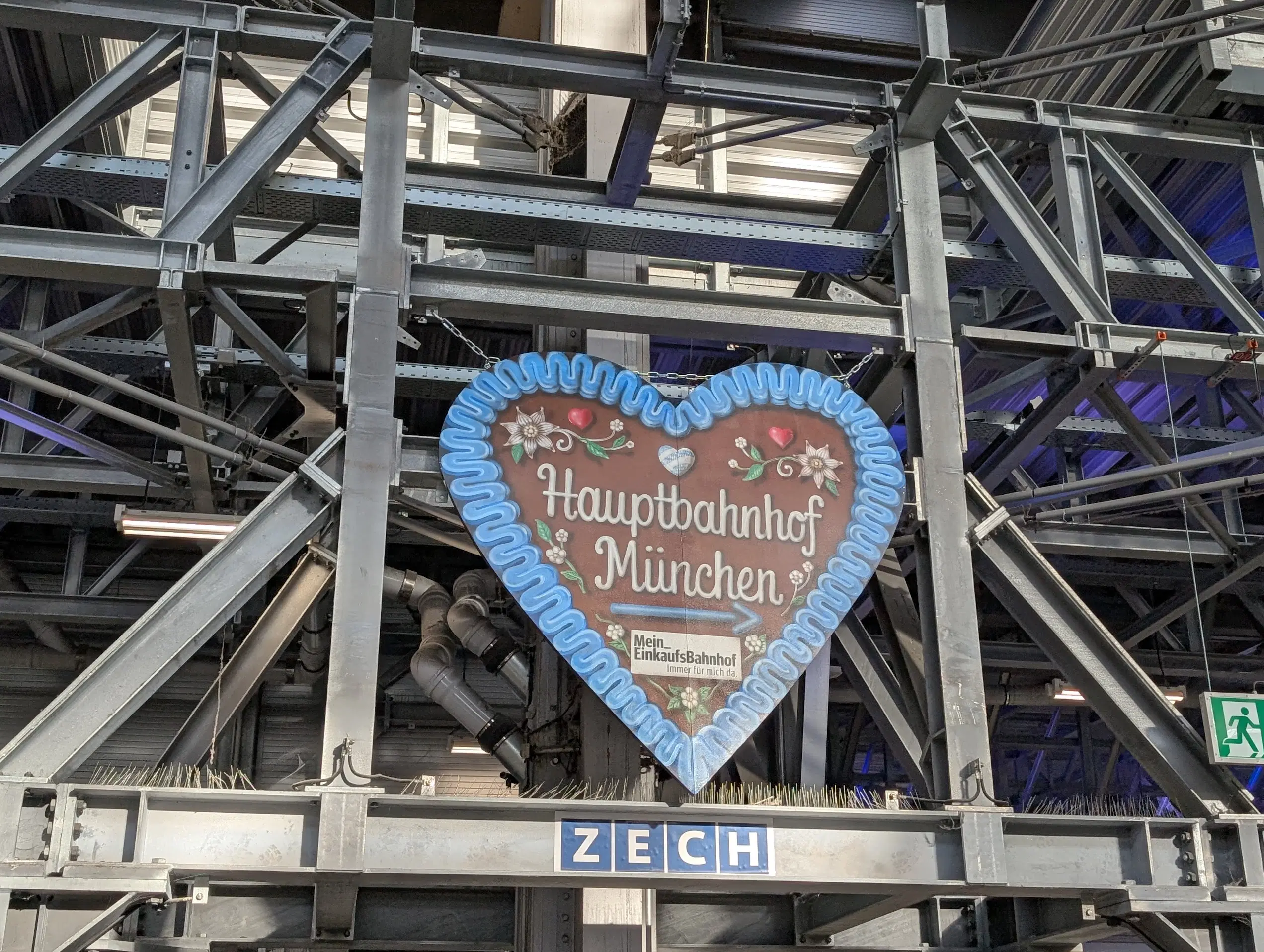 Lebkuchenherz mit der Aufschrift „Hauptbahnhof München“ vor der sichtbaren Stahlkonstruktion des Bahnhofs.